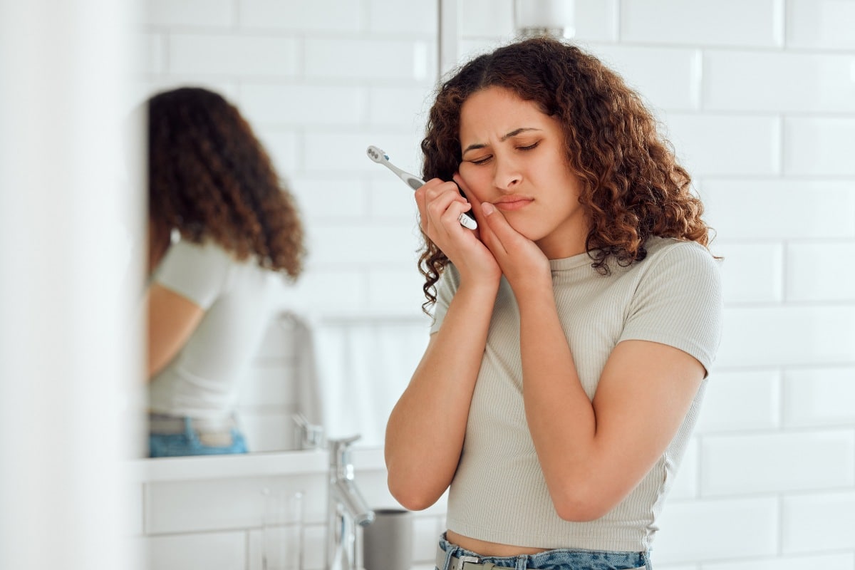 Young woman holding her cheek in pain while brushing her teeth, showing signs of a dental emergency.