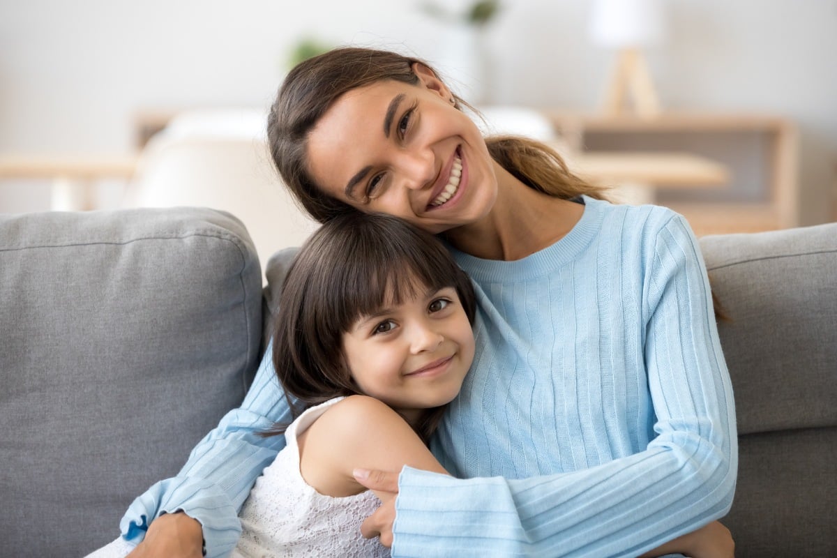 Smiling mother and daughter hugging on a couch, showing healthy, happy smiles.