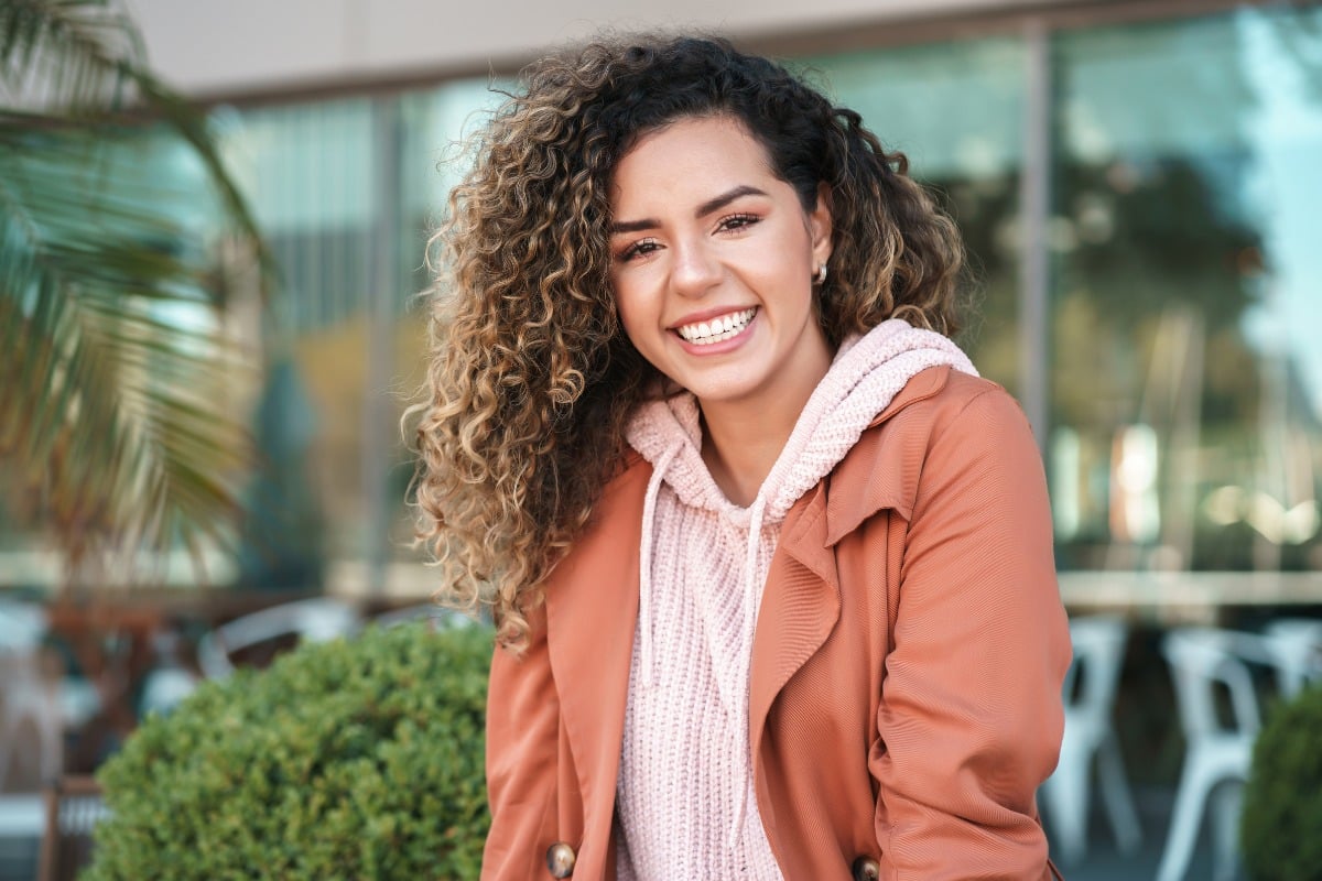 Smiling young woman with curly hair sitting outdoors, wearing a pink hoodie and rust-colored jacket.