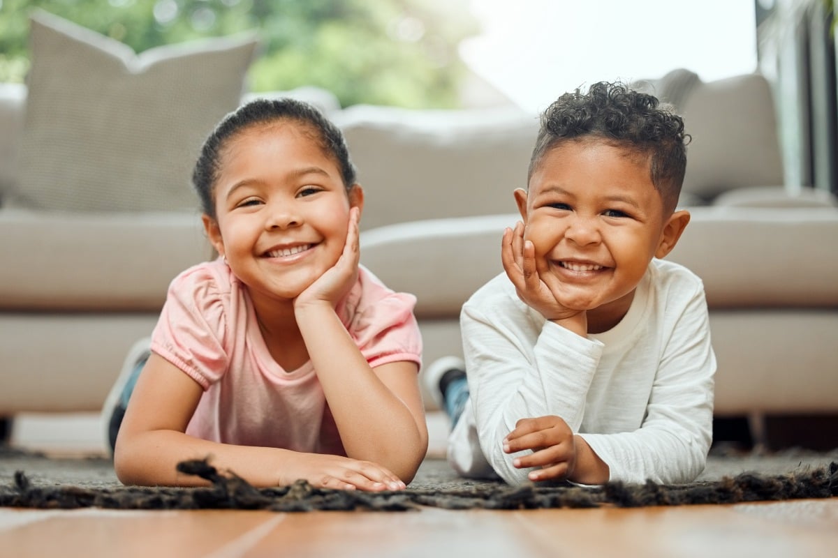 Two smiling young children lying on the floor with their heads resting on their hands.